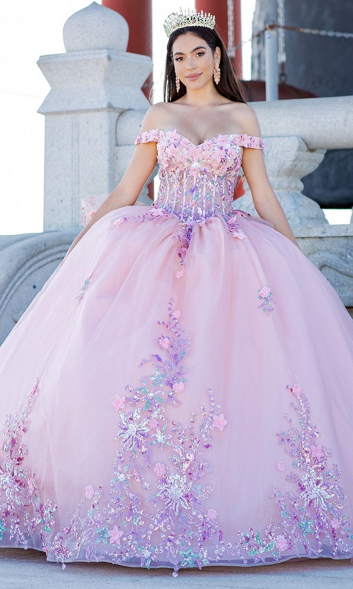 A young woman wearing the Quinceanera Ball Gown 8125J, an off-the-shoulder pink dress with floral embroidery and a tiara, stands outdoors in front of a stone structure.